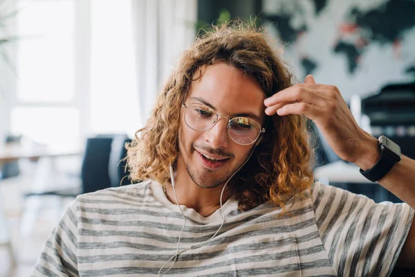 Young curly man in eyeglasses using earphones while working in office