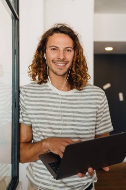Young curly man using laptop and earphones while working in office