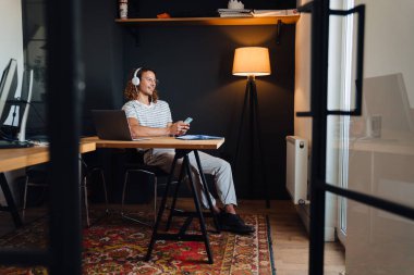 Young curly man using cellphone and headphones while sitting by table in office