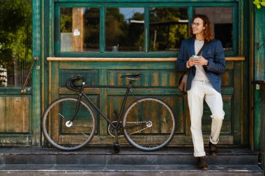 Young smiling ginger man in eyeglasses using smartphone while standing near bicycle on city street