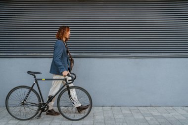 Ginger european man walking with bicycle near grey wall outdoors on city street