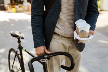 Ginger european man wearing jacket walking with bicycle and coffee on city street