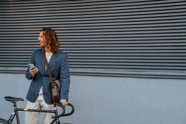 Young ginger white man using mobile phone while standing with bicycle near grey wall outdoors