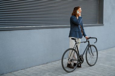 Young ginger joyful man talking on cellphone while standing with bicycle near grey wall outdoors