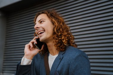 Young smiling ginger man talking on cellphone while standing near grey wall outdoors