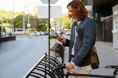 Ginger european man using cellphone while standing with bicycle on city street