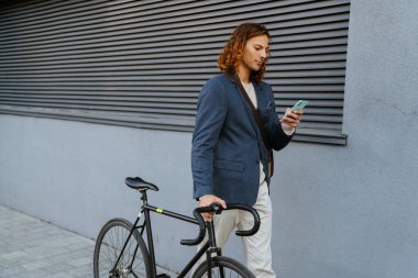 Ginger european man using mobile phone while walking with bicycle on city street