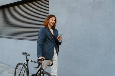 Ginger european man using mobile phone while walking with bicycle on city street