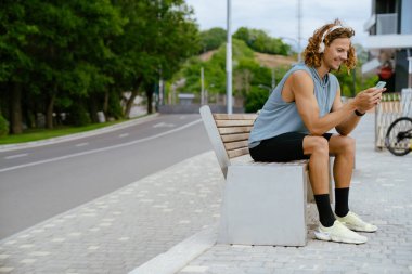 Young handsome smiling sporty long-haired man in headphones with phone sitting on the bench outdoors