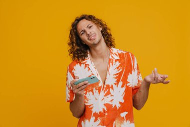 Ginger european man frowning and gesturing while using cellphone isolated over yellow background