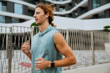 Portrait of young handsome athletic long-haired man in headphones running outdoors