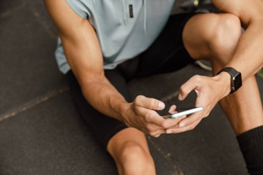 Cropped portrait of young athletic man using phone, while sitting on the ground outdoors