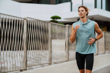 Young handsome athletic long-haired man in headphones running outdoors