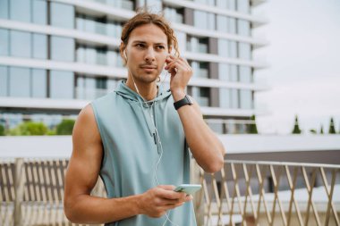 Young long-haired serious handsome man holding phone, putting headphones in ear and looking aside , while standing near modern building outdoors