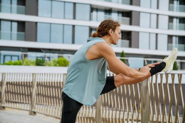 Young athletic long-haired serious handsome man stretching his leg on fence and looking aside , while standing outdoors