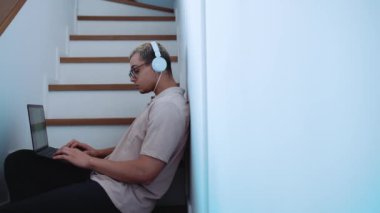 Confident man with piercing and dyed blond hair typing on laptop in headphones on the stairs at home