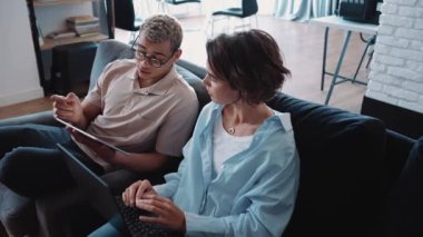 Serious man with piercing and dyed blond hair working on tablet and talking with brunette woman in the office