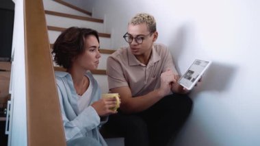 Happy man with piercing and dyed blond hair talking about work on tablet with brunette woman on the stairs