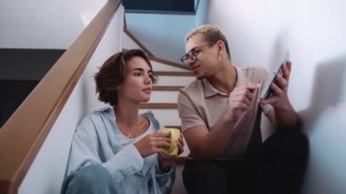 Surprised man with piercing and dyed blond hair talking about work on tablet with brunette woman on the stairs