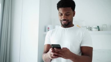 Smiling African man texting on phone while standing in the kitchen at home