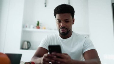 Confident African man texting on phone while sitting in the kitchen at home