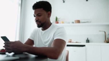 Meditative African man texting on mobile and drinking coffee while sitting in the kitchen at home