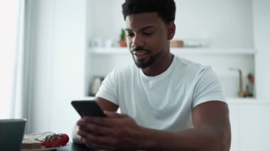 Concentrated African man texting on mobile and drinking coffee while sitting in the kitchen at home