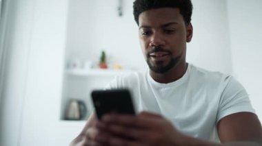 Pensive African man texting on mobile while sitting in the kitchen at home