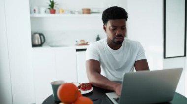 Serious African man working on laptop while sitting in the kitchen at the breakfast