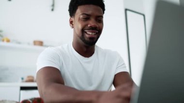 Cheerful African man working on laptop while sitting in the kitchen at home