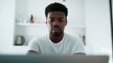 Pensive African man working on laptop while sitting in the kitchen at home