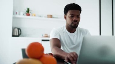 Concentrated African man working on laptop while sitting in the kitchen at the breakfast
