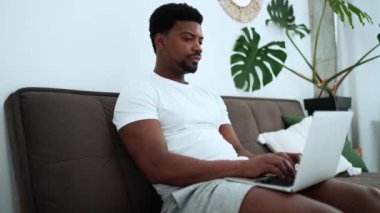 Concentrated African man wearing white t-shirt texting on laptop on the sofa at home