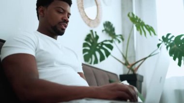 Confident African man wearing white t-shirt texting on laptop on the sofa at home