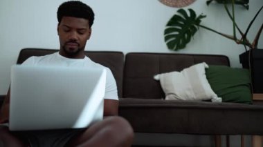 Displeased African man wearing white t-shirt working on laptop while sitting on the floor at home