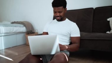 Smiling African man wearing white t-shirt working on laptop on the sofa at home