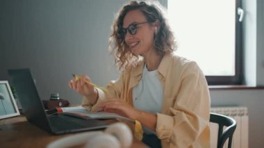 Side view of curly haired cheerful woman talking by video call on laptop at the table at home