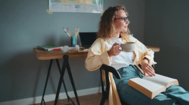 Curly haired positive woman wearing eyeglasses reading book with up of tea at home