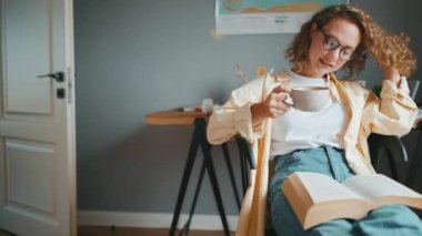 Curly haired happy woman wearing eyeglasses reading book with up of tea at home