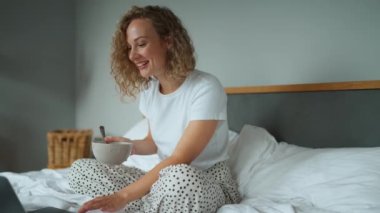 Curly haired laughing woman eating cereal with milk and watching a movie on the laptop on the bed at home