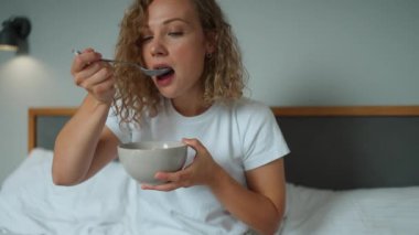 Handsome curly haired woman eating cereal with milk and watching a movie on the laptop on the bed at home