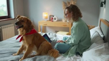 Smiling woman with curly hair typing on laptop while her dog sitting near her on the bed at home