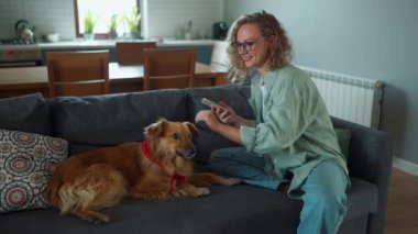 Genç kıvırcık saçlı kadın evdeki kanepede otururken köpeği okşuyor.