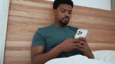 Smiling African brunet man typing on phone while lying on the bed at home
