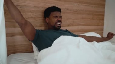 Handsome African brunet man yawning and stretching while lying on the bed at home