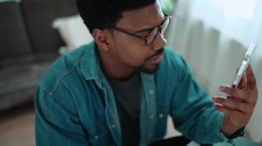 Positive African brunet man talking by video call on mobile while sitting in the room at home