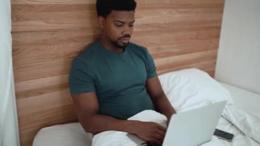 Confident African brunet man texting on laptop while lying on the bed at home