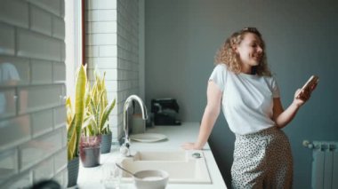 Positive curly haired woman typing on mobile and looking at window in the kitchen at home