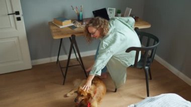 Funny curly haired woman working on laptop and stroking her dog lying on the floor at home