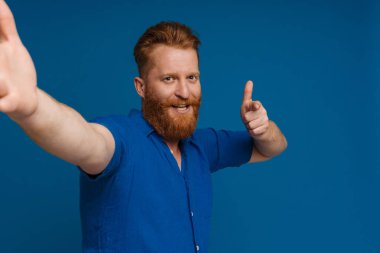 Selfie of adult handsome redhead bearded smiling man doing gun gesture with fingers and looking at camera, while standing over isolated blue background